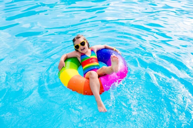 Happy little girl playing with colorful inflatable ring in outdoor swimming pool on hot summer day. Kids learn to swim. Child water toys. Children play in tropical resort. Family beach vacation.